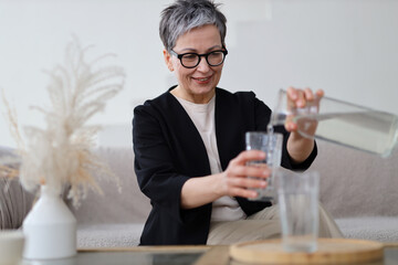 A senior woman smiles happily while holding a glass of water at home, prioritizing her health and...