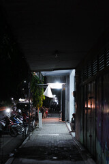 a narrow alleyway at night, with a person sitting on the edge of the sidewalk. The alleyway is dimly lit, and the shadows cast by the buildings and the person create a sense of mystery and intrigue