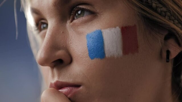 French sports fan closely watches competitions, games, and matches. Close-up of face of young attractive woman with flag of France on her face, whose eyes are carefully looking at something