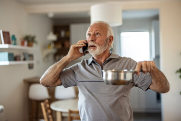 Worried senior man calling plumber while collecting water leaking from ceiling using utensil....