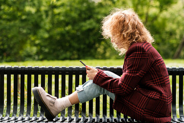 Side view of young woman reading on e-book on a bench park.