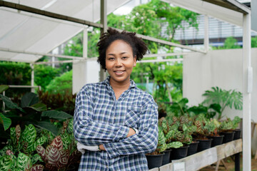 A woman in a plaid shirt stands in front of a greenhouse filled with plants. She is smiling and she is proud of her work