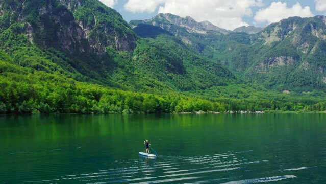 Astonishing Aerial view of Person paddling on SUP board on Mountain Lake in Slovenia. Active Woman does Water Sport on Bohinj alone. Stand up board as popular nature tourism. 4K wide drone shot