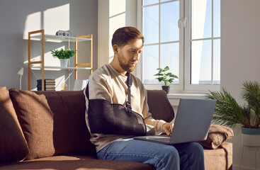 Young man with a broken arm in a medical arm sling working remotely from home, sitting on the...