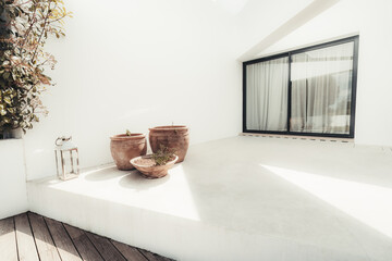 Modern minimalistic patio area with white walls, terracotta plant pots, and a lantern. Large glass sliding door with curtains closed, wooden deck flooring and sunlight casting shadows