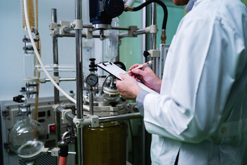 A scientist is writing on a clipboard in front of a lab bench. The clipboard has a pen on it. The scientist is wearing a white lab coat