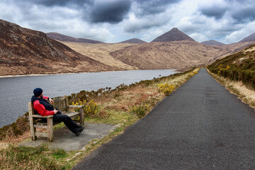 Man in red jacket sit on wood bench lakeshore Silent Valley Reservoir Moure Mountains Lake Northern Ireland UK Europe