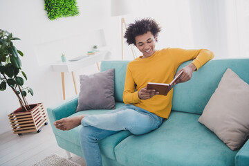 Portrait of nice young man read book sit sofa wear yellow pullover white interior flat indoors