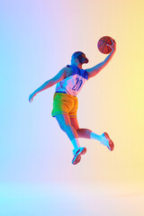 Young athletic woman wearing sports uniform doing perfect slam dunk in motion in neon light against gradient studio background. Concept of professional sport, championship, tournament. Ad