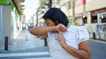 A middle-aged hispanic woman sneezing into her elbow on a busy urban street, signifying health and etiquette awareness. - Powered by Adobe