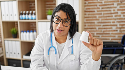 Hispanic woman doctor in glasses holding a medicine bottle in a clinic office with a stethoscope and shelves background