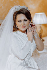 A woman in a white dress is sitting on a bed and putting on her wedding ring. She is wearing a...