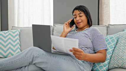 A relaxed hispanic woman discusses paperwork over the phone while sitting with a laptop on a couch indoors.