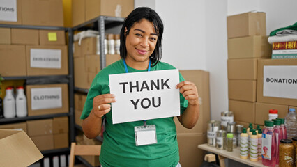 Middle-aged hispanic woman holds 'thank you' sign in a donation center with boxes and supplies.