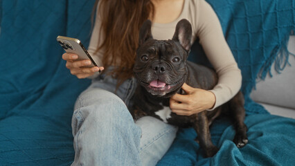 A young hispanic woman sits in her living room holding a cellphone, accompanied by her pet french bulldog on the blue sofa.