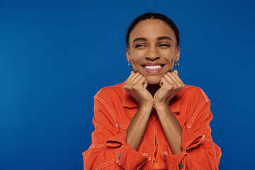 A young African American woman in a vibrant orange outfit smiles, hands on her face, radiating happiness.