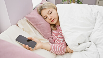 A young blonde woman sleeping in a cozy bedroom, holding a smartphone in her hand, depicting a peaceful indoor scene.