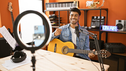 Handsome hispanic man playing guitar in a music studio with on-air sign.