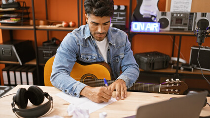 Handsome hispanic man writing music in a recording studio with guitar, laptop, and microphone.