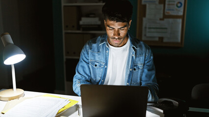 Handsome hispanic man working late on a laptop in a dark office setting, illuminated by desk lamp.