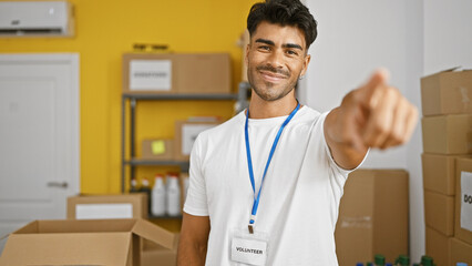 A hispanic man with a beard smiles while volunteering at an indoor warehouse, pointing towards the...