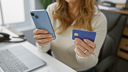 A confident woman holds a credit card and smartphone in an office, symbolizing ecommerce and...