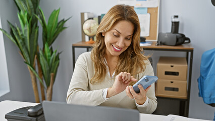 A young hispanic woman smiles while using a smartphone in a modern office setting, reflecting a...