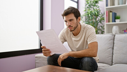 A contemplative young hispanic man reads a document in a bright living room, embodying casual,...