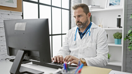 Mature hispanic doctor with grey hair focused on work at computer in a clinic office.
