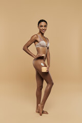 Beautiful African American woman in a bikini applying body cream on a beige background.