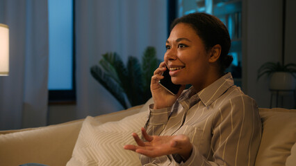 Amazed emotional African American woman ethnic joyful girl talk mobile phone in evening living room...