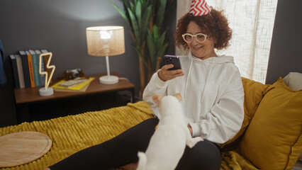 Middle-aged woman in a party hat sitting on a bed with a chihuahua dog, using her smartphone in a cozy bedroom.