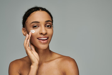 Young African American woman in strapless top delicately applies skincare on cheek with white...