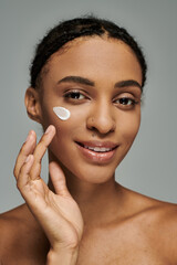 Young African American woman in strapless top applying cream on her face against a grey backdrop.