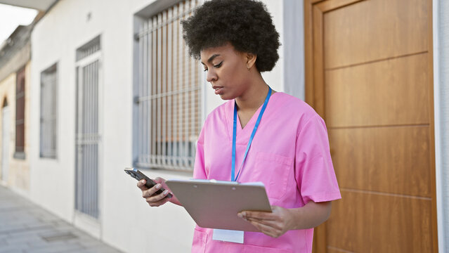African american nurse in pink scrubs holding a tablet and phone outside a clinic - Powered by Adobe