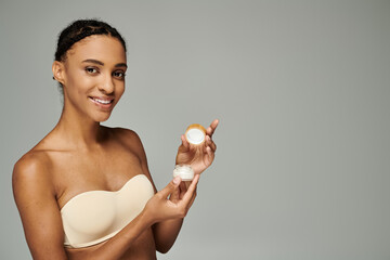 Young African American woman in a bra taking care of her skin, holding a container of cream on a...