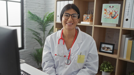 A young, beautiful hispanic woman doctor with brunette hair and glasses smiles warmly in a clinic...