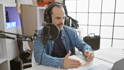 A bearded man wearing headphones speaks into a microphone at a desk with a notepad in a recording...