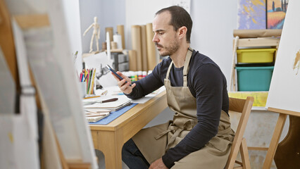 Bald bearded man in studio examining phone with art supplies around.