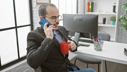 Hispanic businessman in an office on a phone call, holding a coffee mug, wearing glasses, and looking concentrated.