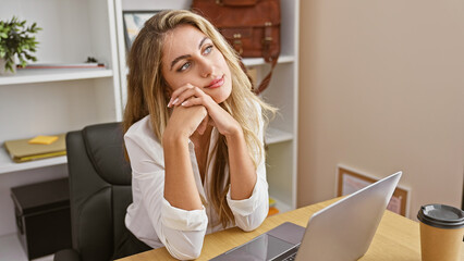 Thoughtful young blonde business worker engrossed in serious work on her laptop in office,...