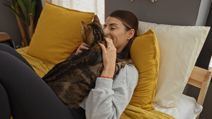Fototapeta premium A young hispanic woman relaxing in bed at home with a kitten, sharing a tender moment in a cozy bedroom with yellow pillows and soft lighting.