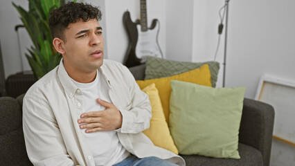 A young man looks uncomfortable while sitting on a couch in a modern living room with a guitar in the background.