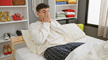 A handsome man in casual attire sitting on a bed in a well-organized bedroom with colorful wardrobe...
