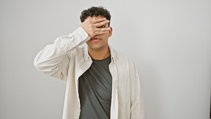 A young adult man with curly hair and beard covering eyes with hand against a white background.
