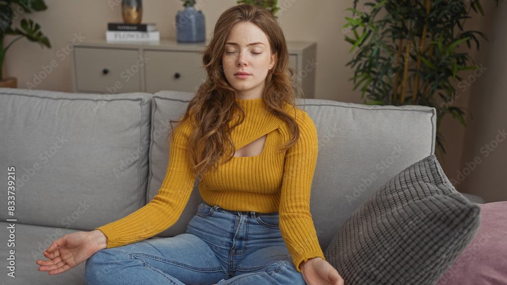 Wall mural A young, attractive blonde woman relaxing in a living room, sitting on a gray couch with eyes closed, wearing a yellow sweater and blue jeans, indoors at home with plants and cushions.