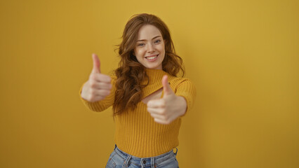 A beautiful young woman with blonde hair gives a thumbs up with both hands while smiling against an...