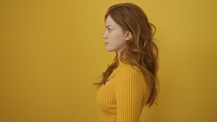 A beautiful young adult woman with blonde hair poses against an isolated yellow background wall,...