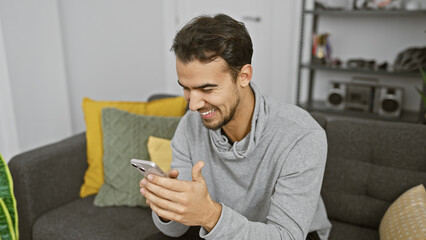 Handsome hispanic man smiling at phone in cozy living room.