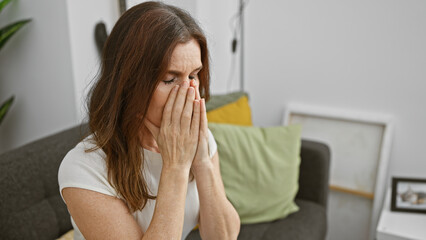 Mature woman in distress with hands covering face sitting on couch at home, conveying emotion of...
